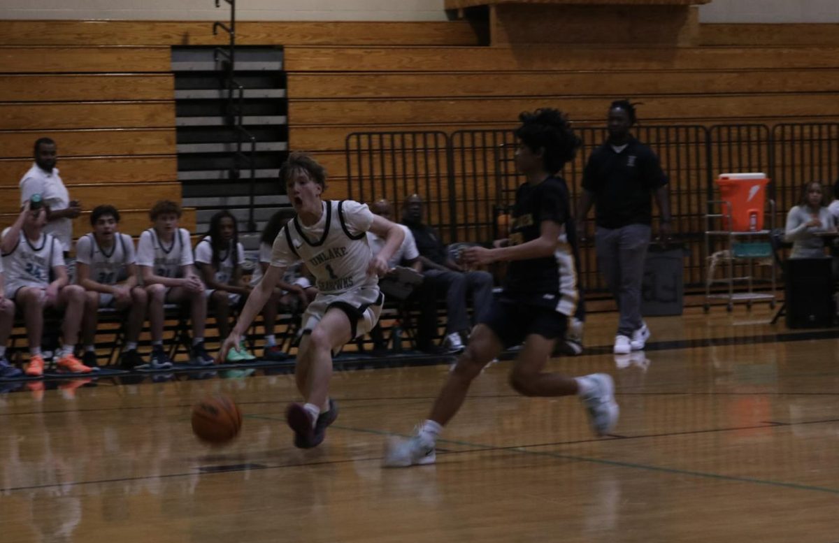 Aaron Hanstedt; Sophomore, is dribbling the ball quickly down the court, leaning forward as he pushes the ball ahead with one hand. His teammates cheer him on from the sidelines.